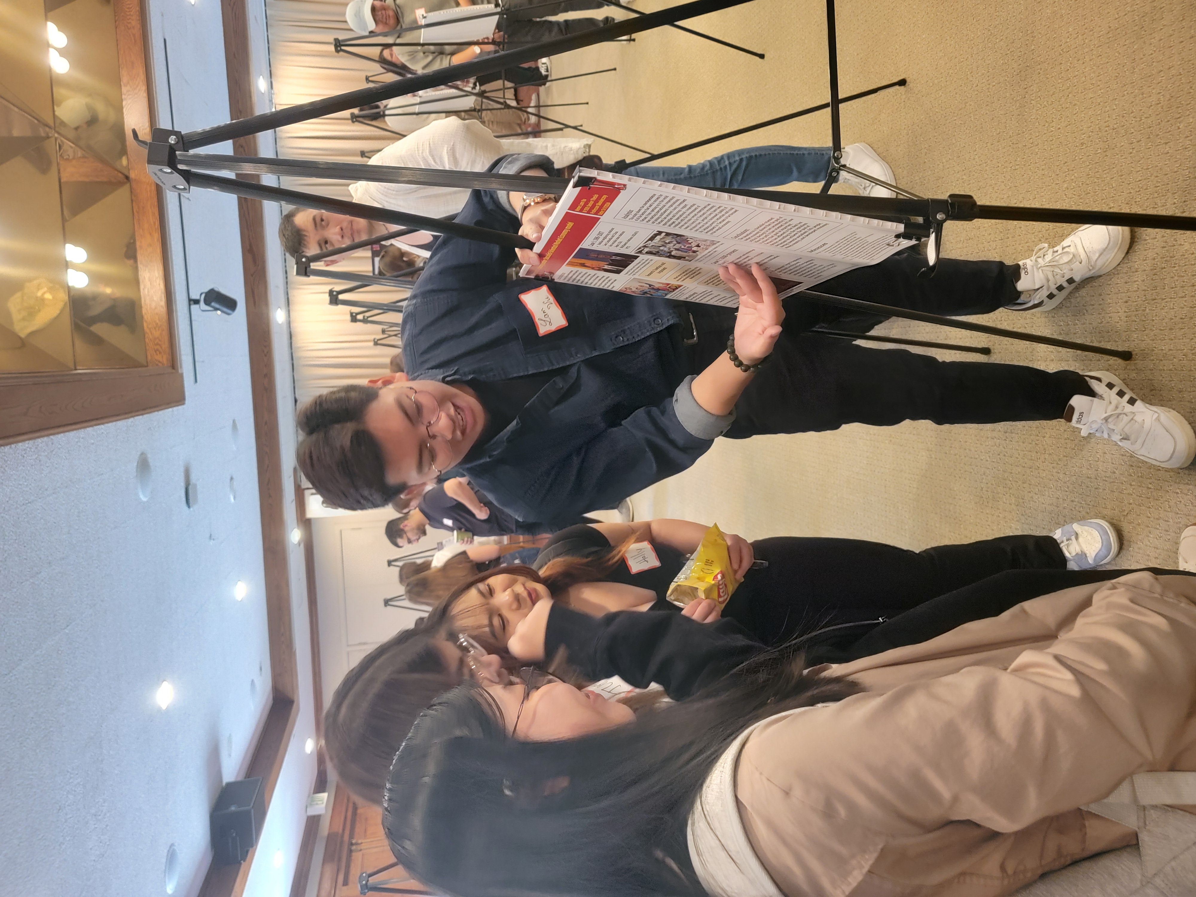 A student from IR 550 faces his research poster, gesturing and explaining his work to another three students, who cluster around him and watch intently.