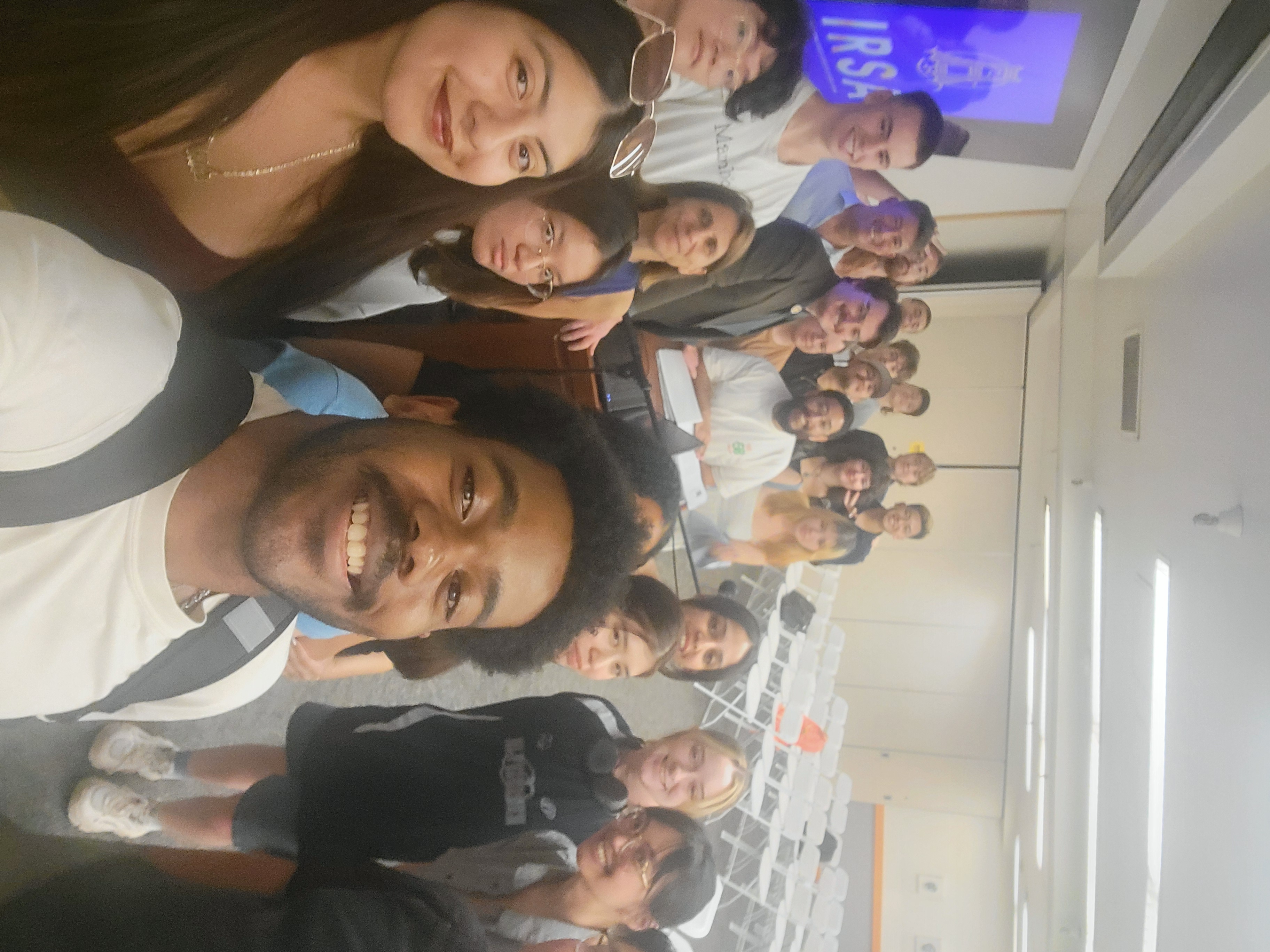 22 students gather around Dan Buschmeyer and Professor Martin Morales, standing near a podium, for a selfie. All are smiling at the camera.