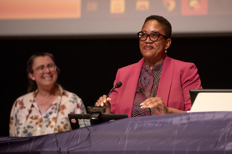 Ifeoma Nwankwo smiles and gestures behind a podium. She is wearing a pink blazer, a spotted dress with a high collar, glasses, and pearl earrings. Burcu Ellis smiles and looks towards her in the background.