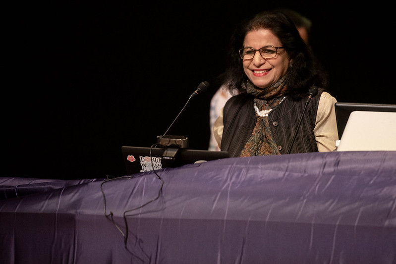 Nandini Tandon smiles from behind a podium. She is wearing glasses, a pearl necklace, and a floral shirt.